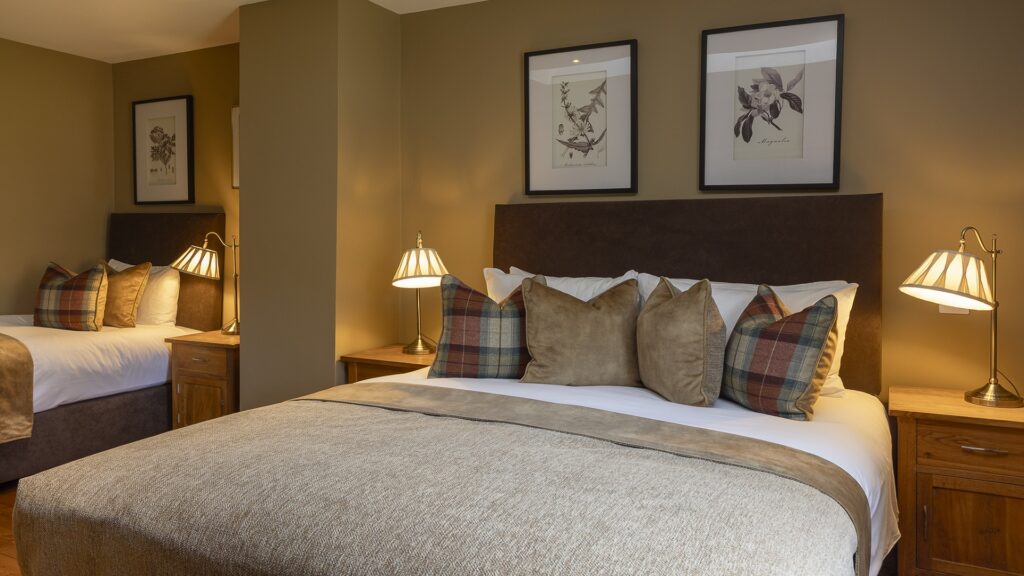 A hotel room with two neatly made beds, each with plaid and neutral pillows, wooden nightstands, lamps, and framed botanical prints on the wall.