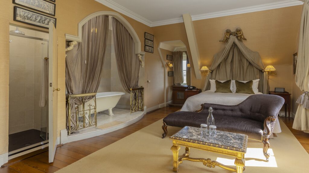 A spacious bedroom with a canopy bed, a black velvet sofa, a marble coffee table, and a bathroom area featuring a clawfoot tub and a separate shower.