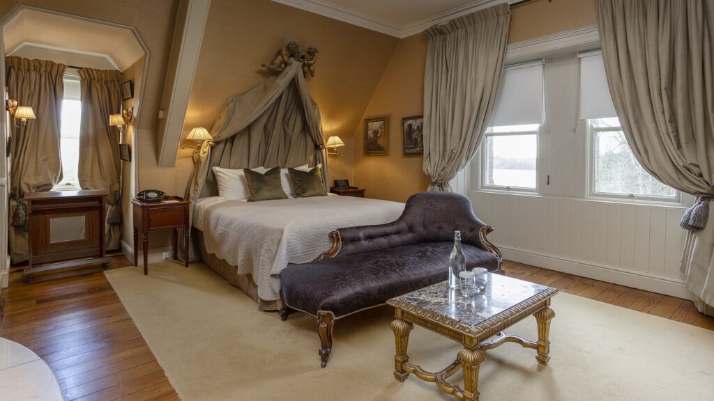 A well-lit bedroom with a double bed, ornate headboard drapes, antique furniture, a small sofa, and a window overlooking a snowy landscape.