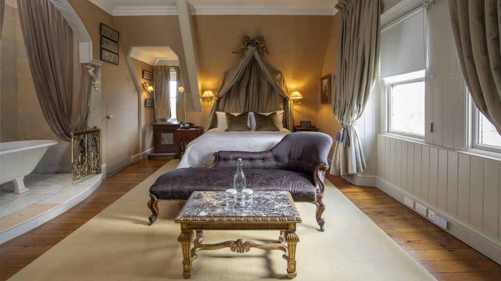 An elegant bedroom features a canopy bed, a velvet chaise lounge, a marble-topped coffee table, and a freestanding bathtub, with warm lighting and large curtained windows.