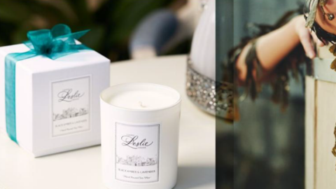 A white candle labeled “Leslie” sits on a table next to a matching white gift box with a teal ribbon.