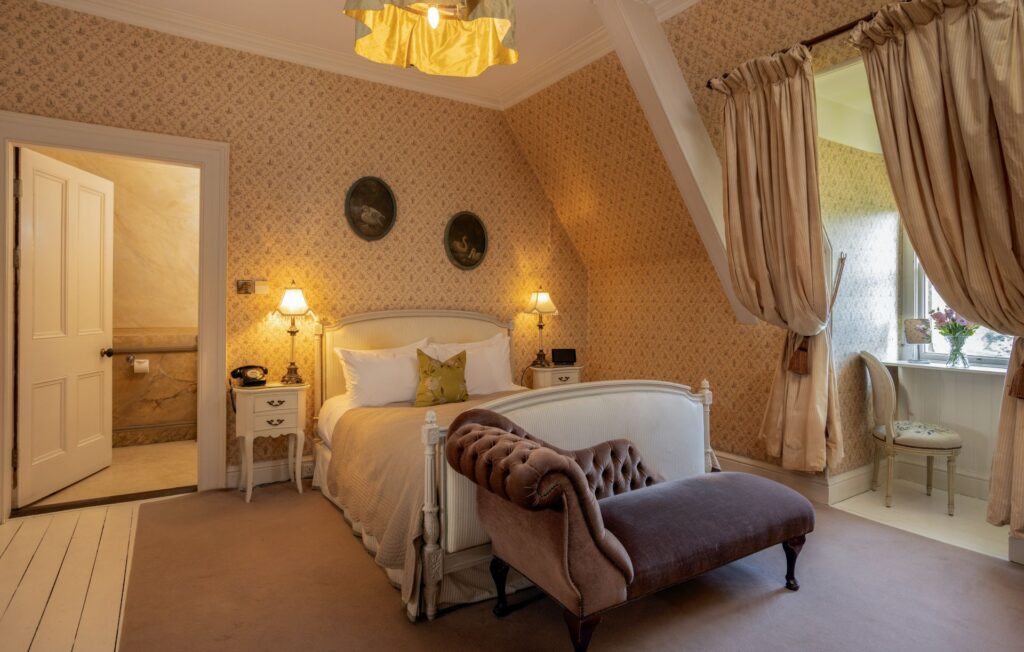 A vintage-style bedroom with a double bed, cushioned bench, two lamps, patterned wallpaper, en suite bathroom, and a window desk with beige drapes and a chair.
