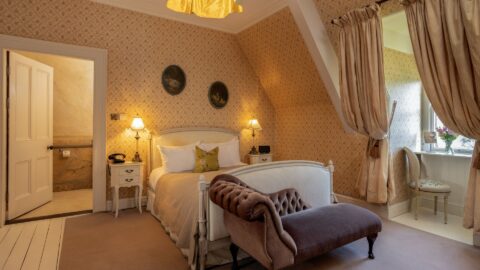 A vintage-style bedroom with a double bed, cushioned bench, two lamps, patterned wallpaper, en suite bathroom, and a window desk with beige drapes and a chair.