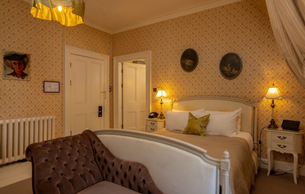 Governess 2 A warmly lit bedroom with vintage wallpaper, a double bed, two bedside tables with lamps, a brown sofa, and decorative wall art.
