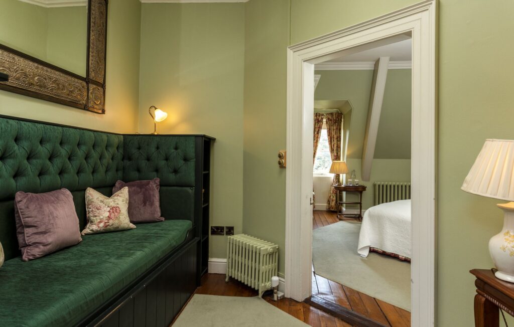 Lionels 1 A room with a green velvet daybed, pillows, and lamp leads to a bedroom with a bed, side table, radiator, lamp, and window with patterned curtains.