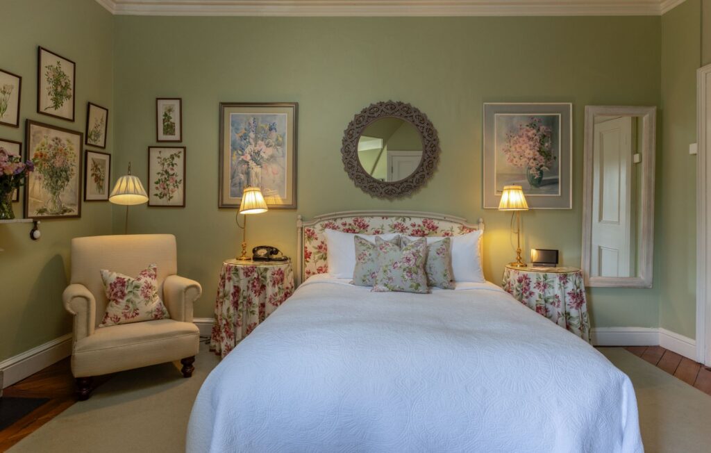 Lionels 3 A neatly made bed with a white cover is centered in a room with green walls, floral decor, an armchair, two lamps, framed art, and a round mirror above the headboard.