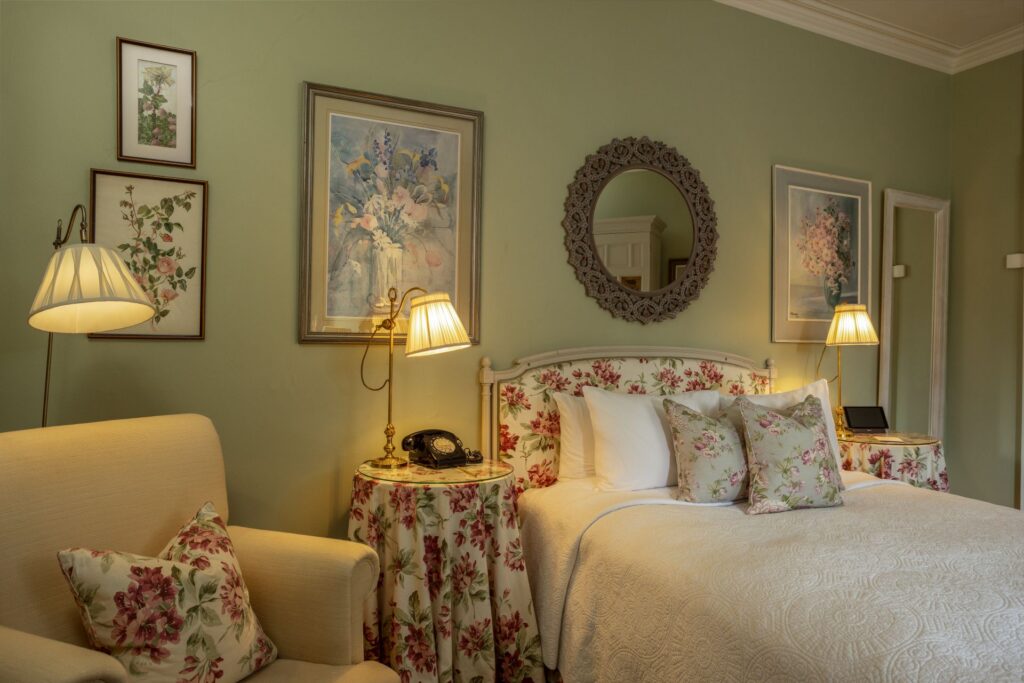 Lionels 4 A cozy bedroom with a floral theme, featuring a bed, armchair, floral pillows, lamps, framed art, and a round mirror on light green walls.