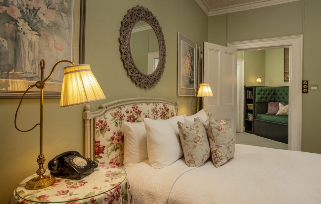 A traditional bedroom with sage green walls, a floral upholstered headboard, white bedding, brass lamps, an oval mirror, and a vintage rotary phone on the bedside table.