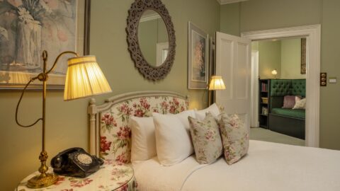 A traditional bedroom with sage green walls, a floral upholstered headboard, white bedding, brass lamps, an oval mirror, and a vintage rotary phone on the bedside table.