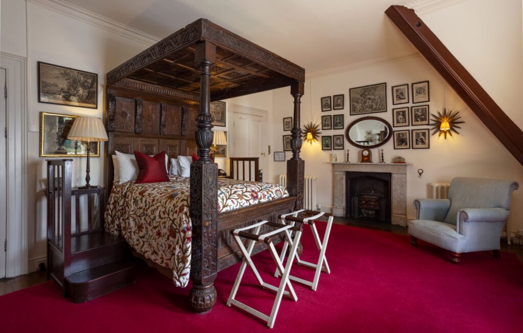 Normans 1 Room with a carved wooden four-poster bed, red carpet, fireplace, gray armchair, and walls decorated with framed art and photographs.