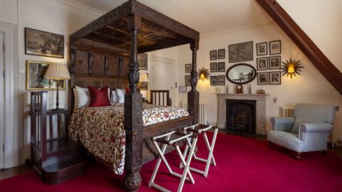 Room with a carved wooden four-poster bed, red carpet, fireplace, gray armchair, and walls decorated with framed art and photographs.