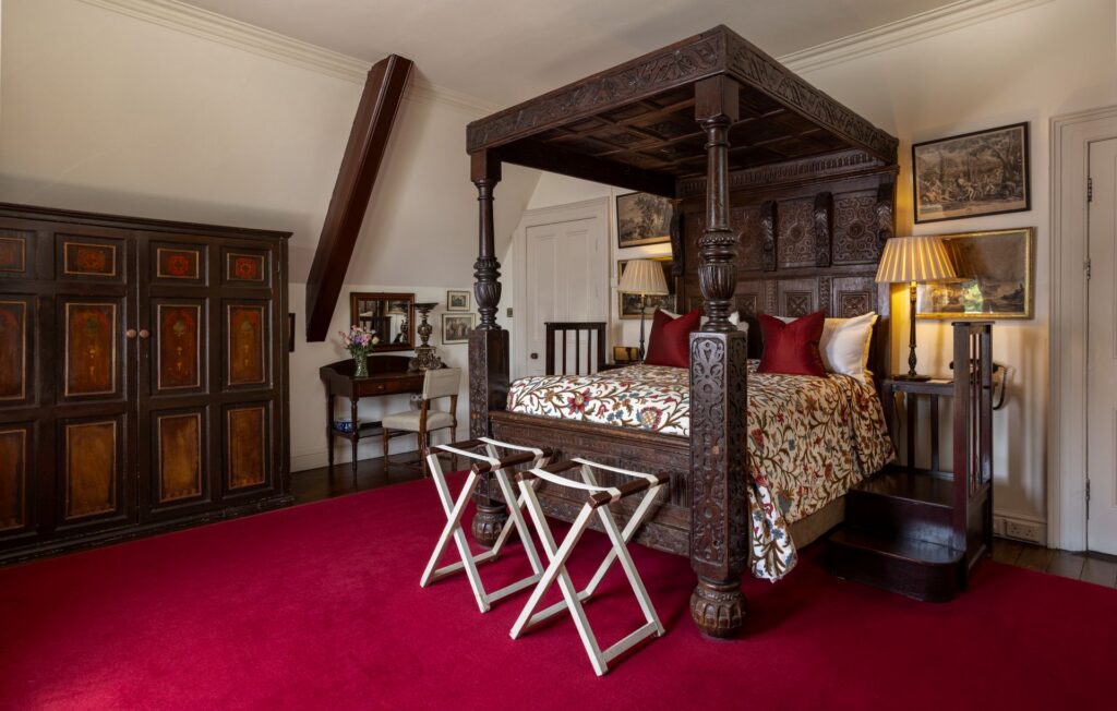 Normans 2 Room with a large, carved wooden canopy bed, red carpet, two folding benches, an ornate wardrobe, a writing desk with a mirror, lamps, and framed art on cream-colored walls.