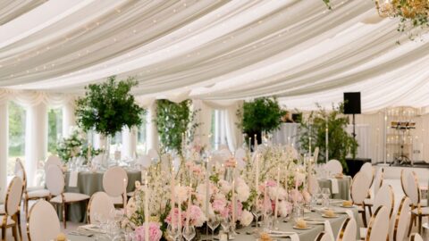 Elegant banquet hall with draped ceiling, chandeliers, and tables set with floral centerpieces and white chairs. Lush greenery and soft lighting create a sophisticated atmosphere.