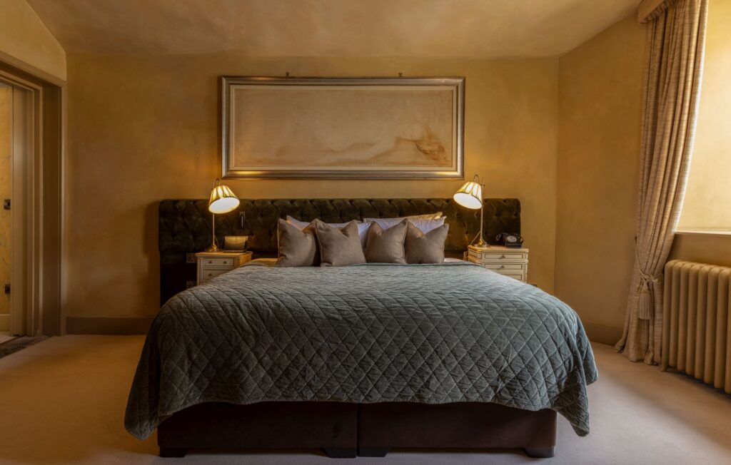 ROC1 A neatly made bed with four pillows and a quilted cover stands between two nightstands with lamps in a softly lit beige bedroom. A large framed artwork hangs above the headboard.
