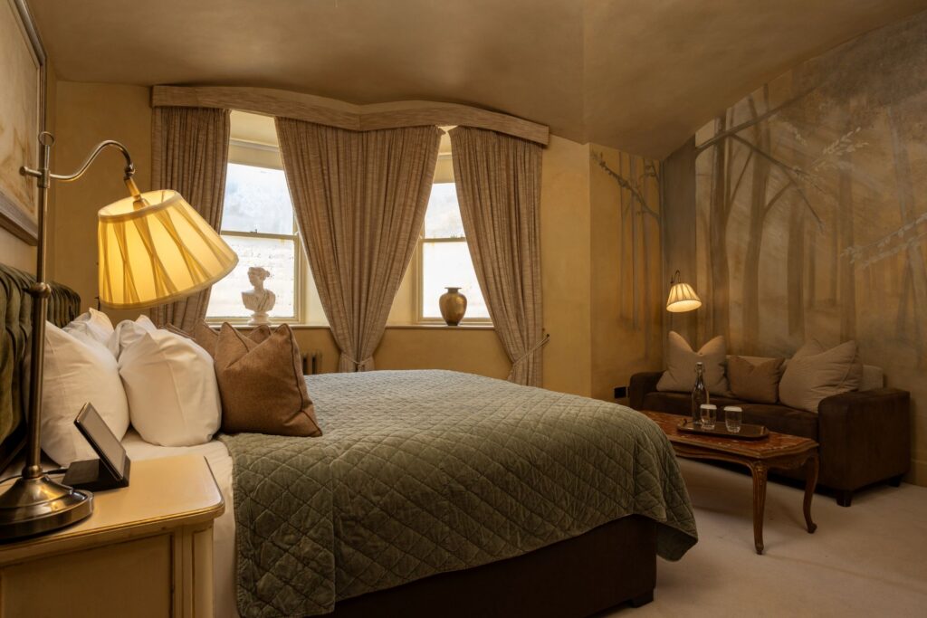 A cozy bedroom with a large bed, brown sofa, two lamps, and a mural of trees on the wall; three windows with beige curtains let in natural light.