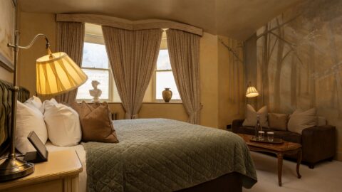 A cozy bedroom with a large bed, brown sofa, two lamps, and a mural of trees on the wall; three windows with beige curtains let in natural light.