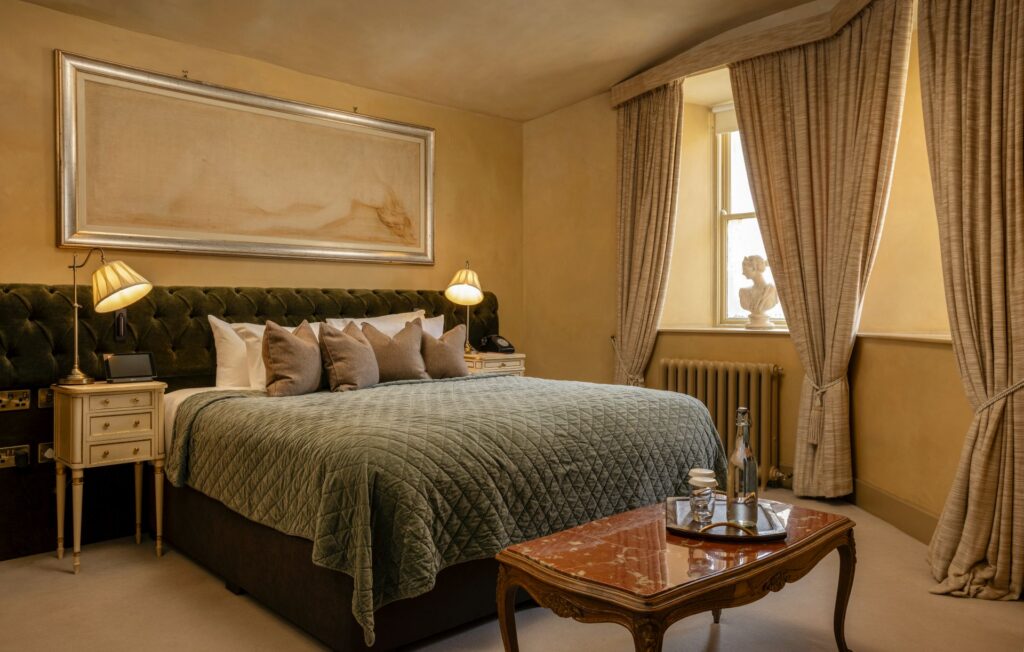 Elegant bedroom with a green tufted bed, marble-top table, two lamps, beige curtains, and a sculpture by the window. Neutral tones dominate the decor.