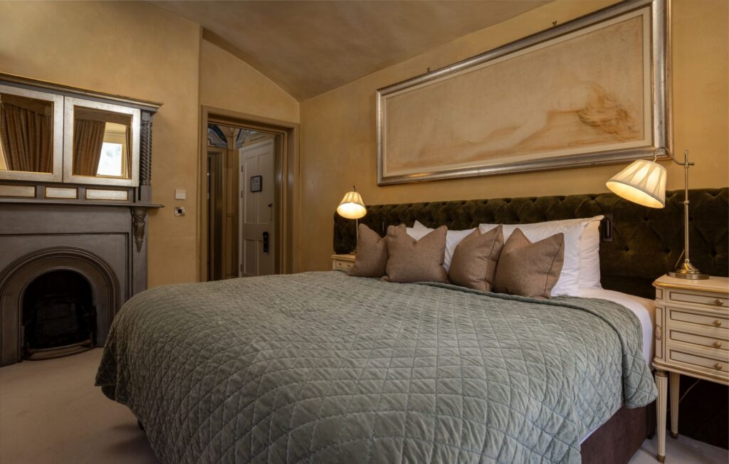 ROC4 A neatly made bed with green quilt and brown pillows, flanked by lamps and nightstands, in a warmly lit bedroom with a fireplace and large framed artwork above the headboard.
