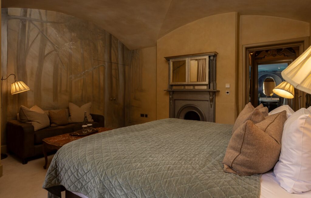 ROC5 A cozy bedroom with a large bed, cushioned sofa, wall mural of trees, fireplace, and warm lighting. A mirror reflects an adjoining room.