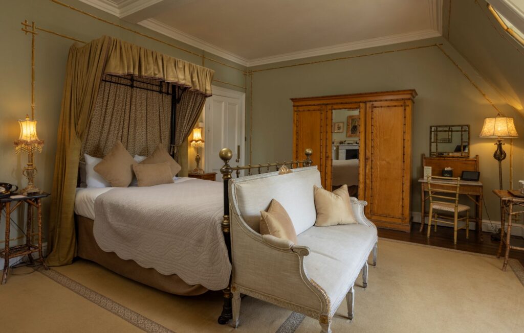 Elegant bedroom with a canopy bed, cushioned bench, wooden wardrobe, and dressing table. Warm lighting and neutral tones create a cozy, classic atmosphere.