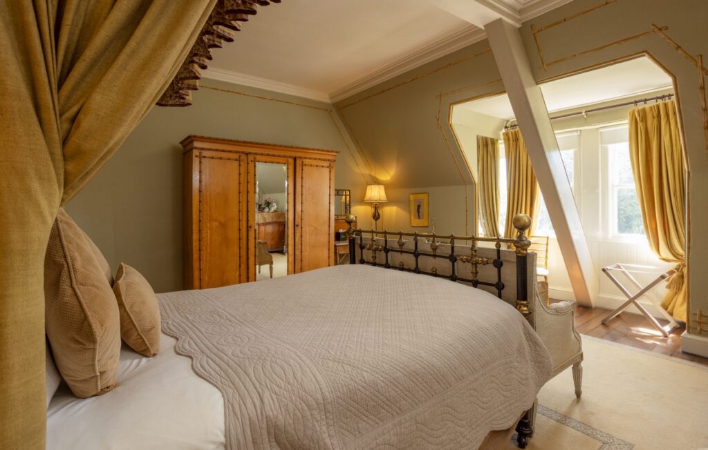 Bright bedroom with a metal bed frame, beige bedding, wooden wardrobe, lamp, and large curtained windows letting in natural light.