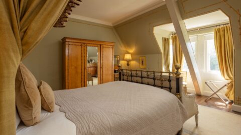 Bright bedroom with a metal bed frame, beige bedding, wooden wardrobe, lamp, and large curtained windows letting in natural light.