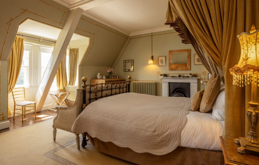 Seymours 3 A cozy bedroom with a large bed, elegant gold details, draped curtains, a fireplace, vintage decor, and natural light streaming through a tall window.