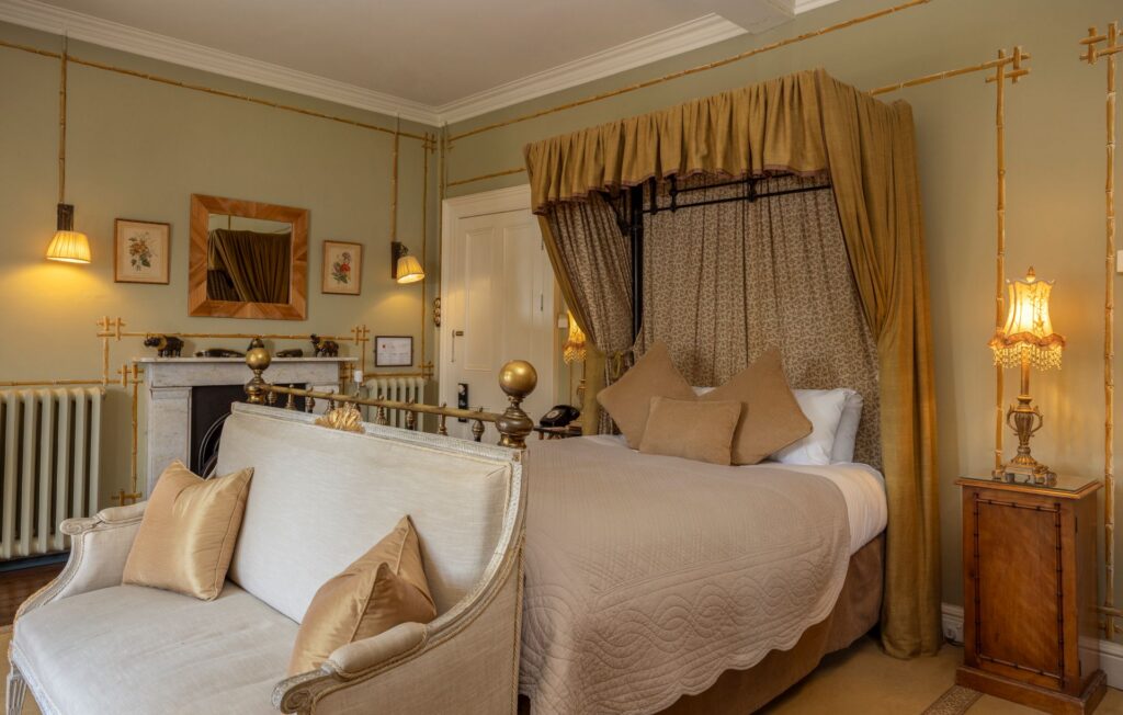Seymours 4 Elegant bedroom with a canopy bed, beige bedding, gold accents, a small sofa at the foot of the bed, vintage lamps, framed art, and a marble fireplace.