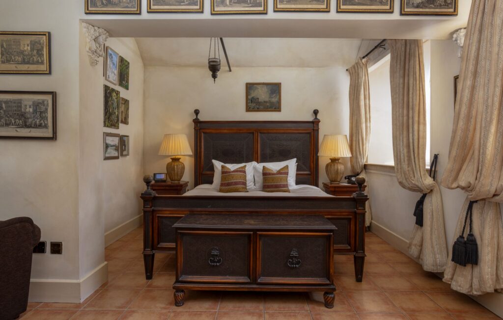 Sir Johns 1 A traditional bedroom with a wooden bed, two lamps on bedside tables, framed artwork on the walls, and large windows with beige curtains.