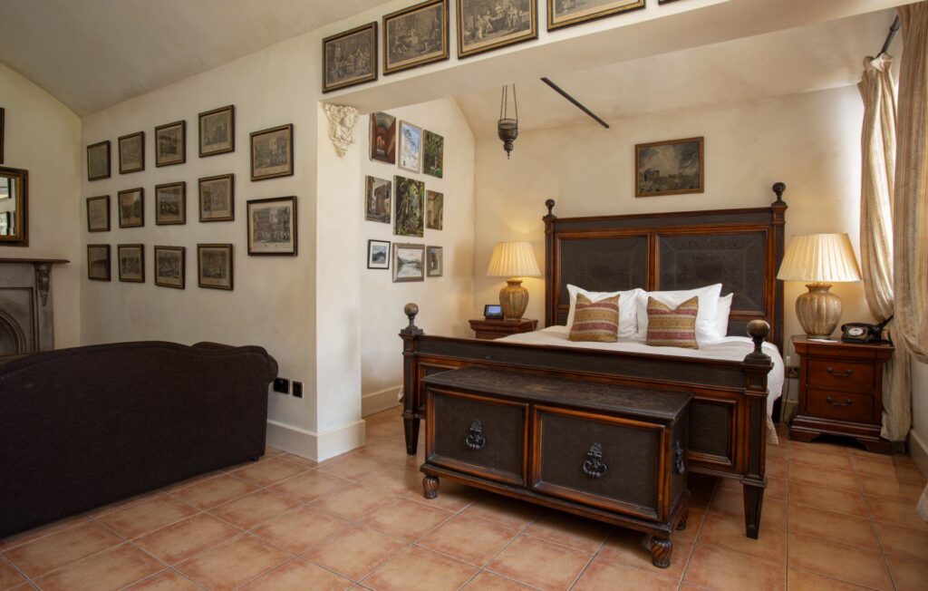 A bedroom with a wooden bed, side tables with lamps, a brown sofa, and framed pictures on cream-colored walls above tiled flooring.