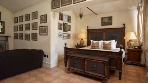 A bedroom with a wooden bed, side tables with lamps, a brown sofa, and framed pictures on cream-colored walls above tiled flooring.