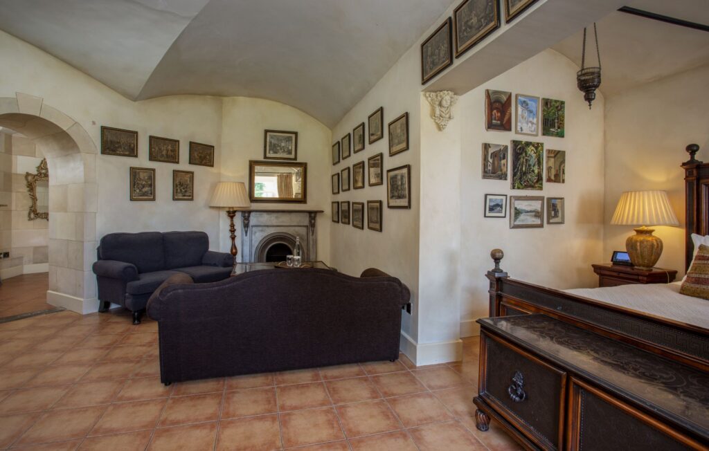 Sir Johns 3 A vintage-style room with tiled floor, dark sofa, armchair, fireplace, framed pictures on cream walls, wooden bed, and lamp on a side table.