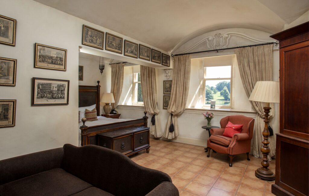 Sir Johns 4 A traditional room with framed artworks on white walls, a bed, wooden furniture, a brown armchair with a red pillow, large windows, and tiled flooring.