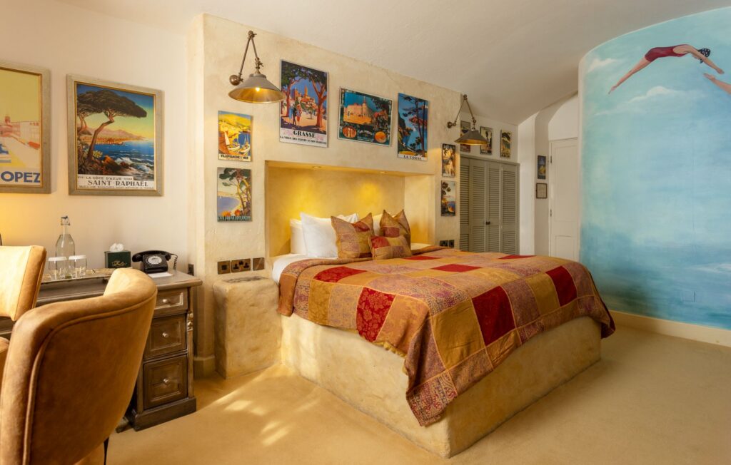 Cozy bedroom with a patchwork quilt, vintage travel posters on the wall, a mural of a swimmer, a wooden desk, and warm ambient lighting.
