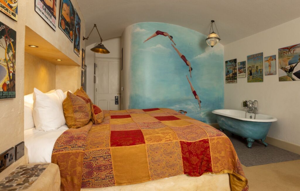A cozy bedroom with a patterned bedspread, vintage posters on the walls, a mural of divers, and a freestanding blue bathtub.