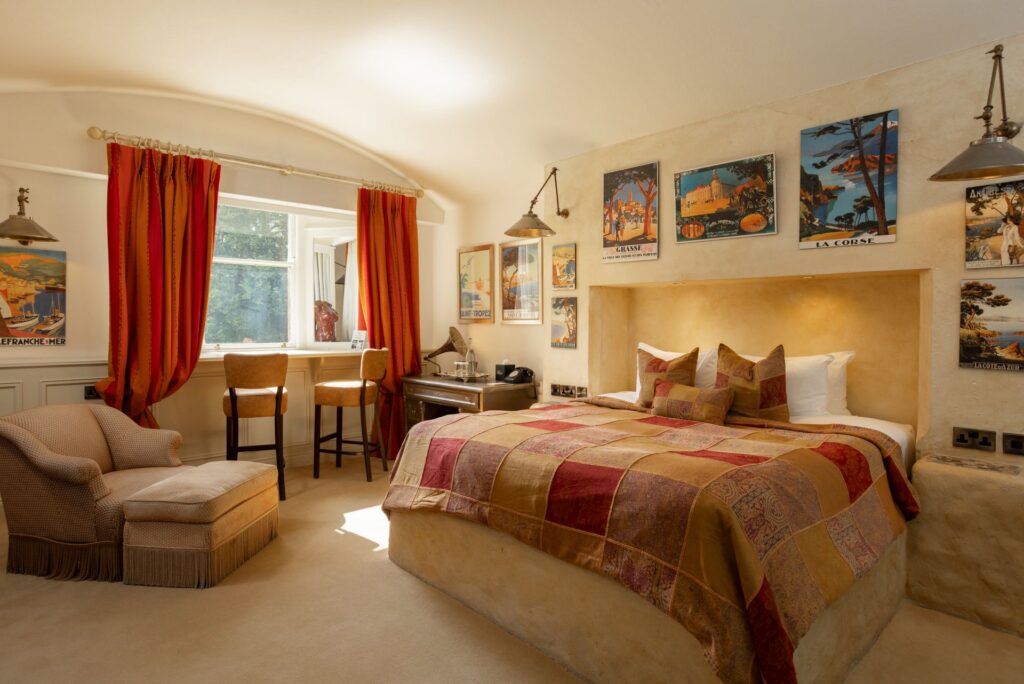 Well-lit bedroom with a large bed, colorful patchwork bedding, armchair, desk by window, and vintage travel posters on the walls.