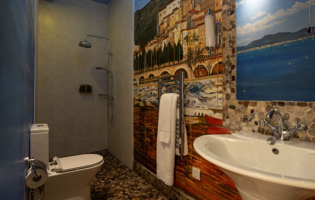 A bathroom with a toilet, open shower, towel rack, sink, and wall art depicting a coastal town and sea view.