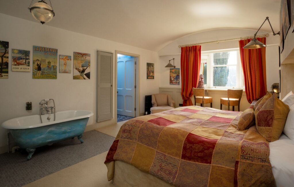 A bedroom with a patchwork bedspread, vintage posters, freestanding blue bathtub, two chairs by a window with red curtains, and soft lighting.