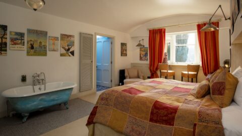 A bedroom with a patchwork bedspread, vintage posters, freestanding blue bathtub, two chairs by a window with red curtains, and soft lighting.