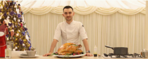 A chef standing behind a kitchen island with a roasted turkey on a platter, next to a decorated Christmas tree.